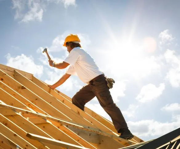 A construction worker in a hard hat and white shirt is hammering nails into wooden beams of a roof structure under the bright sun.