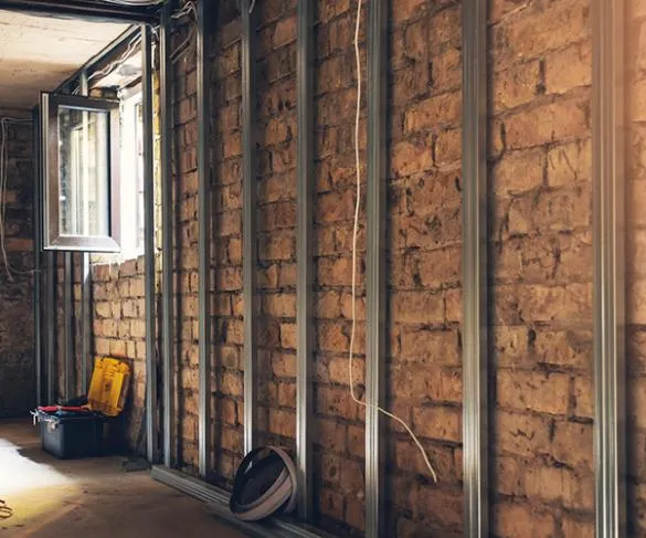 A room under construction with exposed brick walls and metal studs, an open window, a tool box, and an extension cord on the floor.