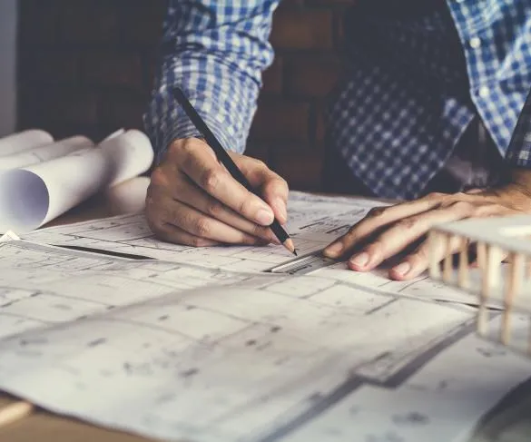 Person in a checkered shirt drawing architectural plans on a large sheet of paper, with rolled-up blueprints on the table.