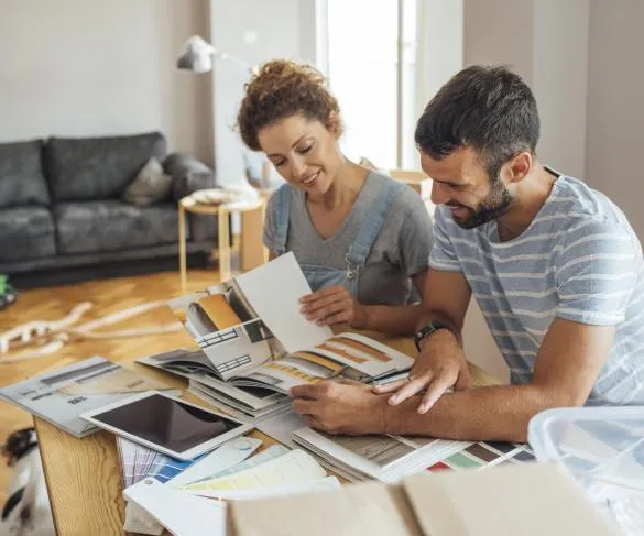 A man and woman sit at a table looking at color swatches and design magazines, planning home decor. Various papers and tablets are on the table.