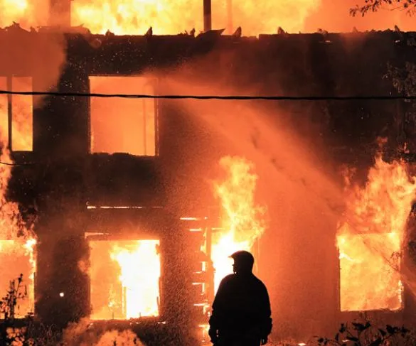 A firefighter stands in front of a burning building at night, with flames coming out of the windows and the structure engulfed in fire.