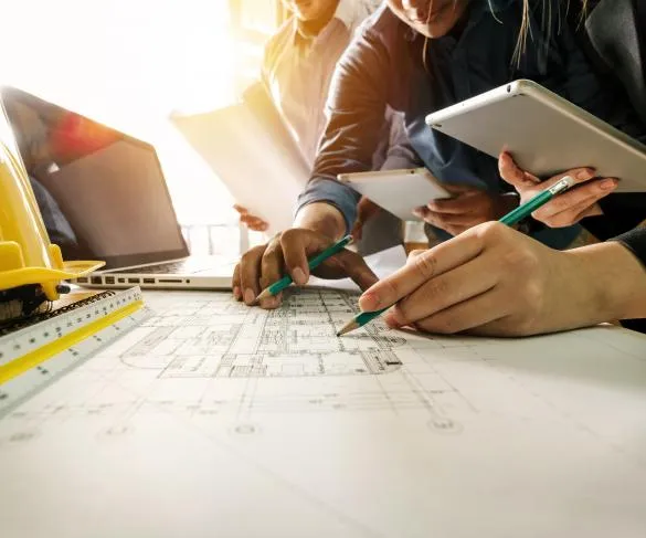 People collaborating on architectural blueprints, with laptops, tablets, and construction helmets visible on the table.