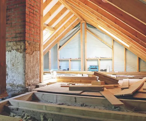 An attic under construction with exposed wooden beams, a brick wall, and an assortment of wood pieces scattered on the floor.