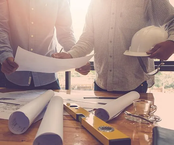 Two people reviewing blueprints on a table with rolled plans, a level, and a helmet in a bright room.