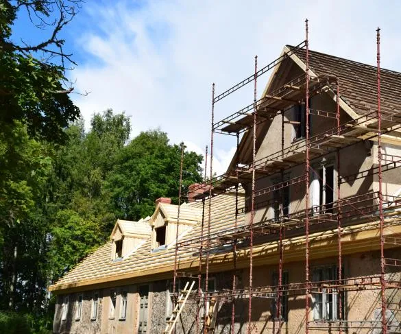 A large house under construction with scaffolding around it, surrounded by trees on a clear, sunny day.