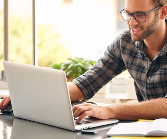 A man wearing glasses and a plaid shirt works on a laptop at a desk, with a smartphone, notebook, and potted plant nearby.