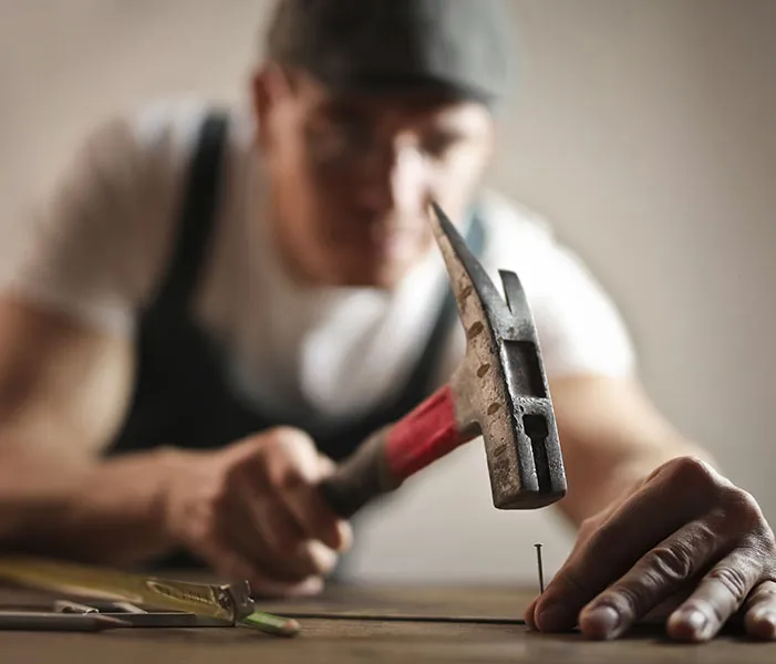 Person hammering a nail into a surface, focusing intently on the task. Various tools are visible nearby.