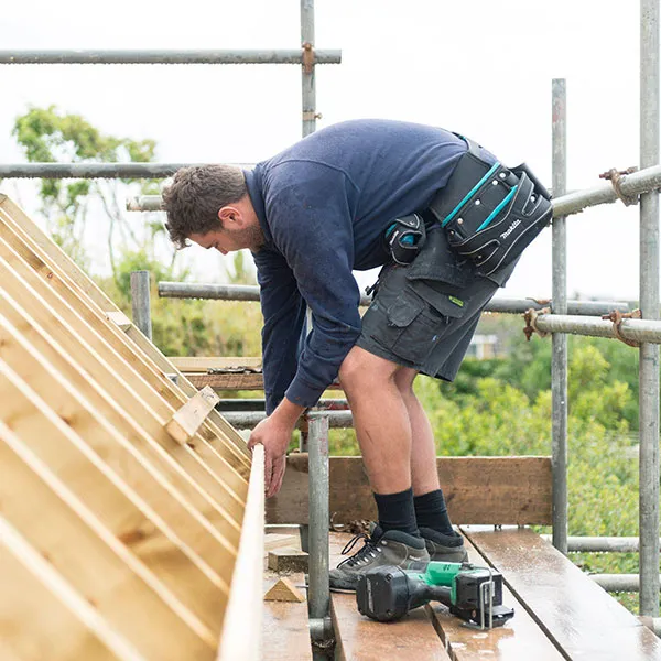 A construction worker on a scaffold, attaching wooden beams to a roof frame with various tools nearby.