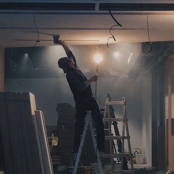 Person on a ladder sanding a ceiling with a power tool in a dimly lit room under construction, holding a light bulb in the other hand.