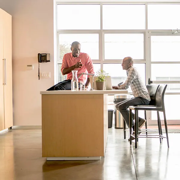 Two people are conversing in a modern kitchen with an island and large windows. One is standing, and the other is seated on a bar stool.