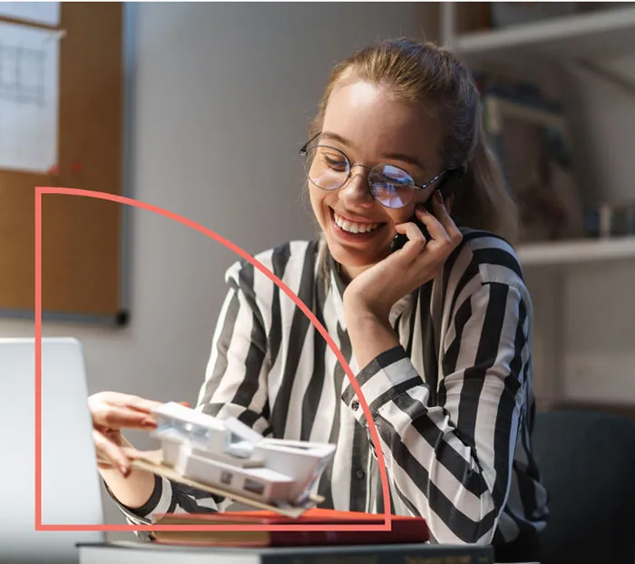 A person wearing glasses and a striped shirt talks on the phone, smiling, while holding a model house and sitting at a desk with a laptop.