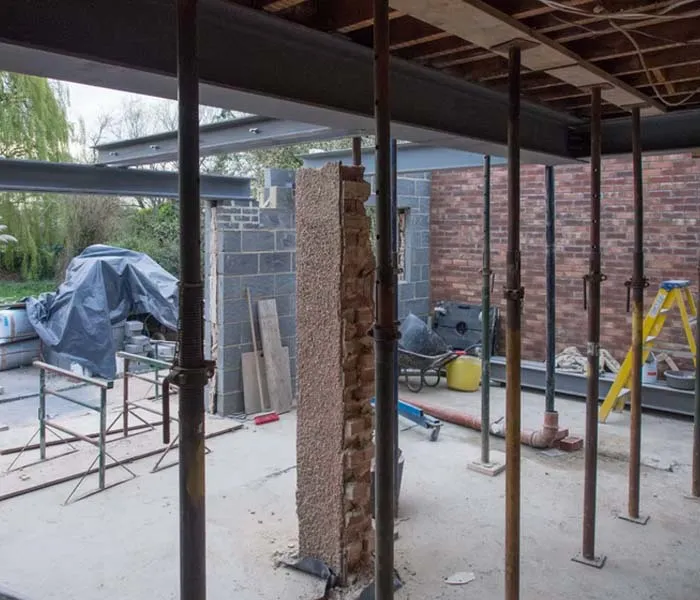 Construction site with steel supports, a partially demolished wall, and building materials scattered around. Ladder stands in the corner.