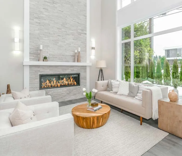 Modern living room with a light color palette, featuring a stone fireplace, white furniture, a wooden coffee table, and large windows.