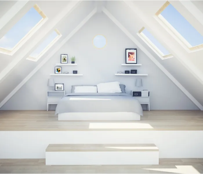 A minimalist attic bedroom with a double bed, skylights, and framed pictures on shelves within a white-walled space.