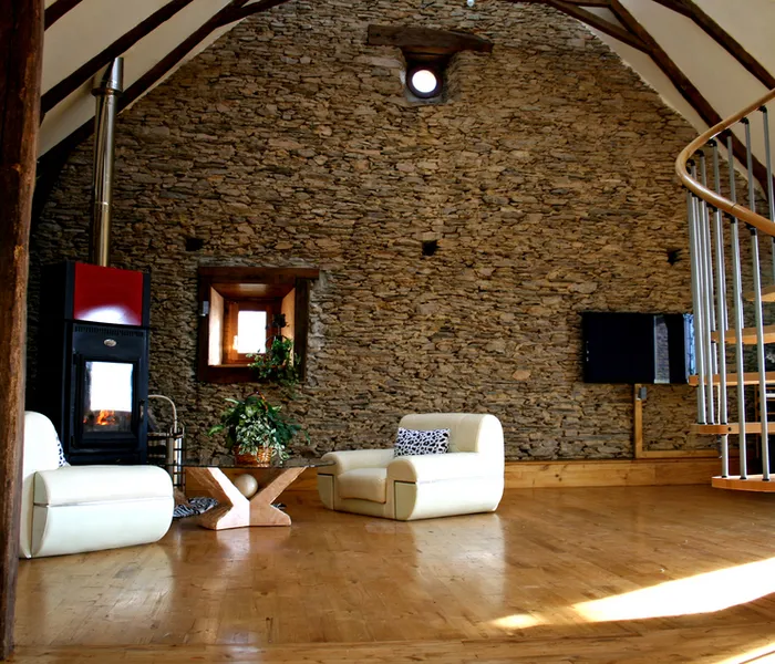 A rustic living room features stone walls, wooden floors, a spiral staircase, two white chairs, a fireplace, and a TV.