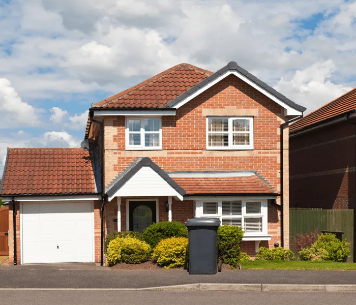 A two-story brick house with a brown tiled roof, garage, and front garden. A black wheelie bin is placed near the driveway.