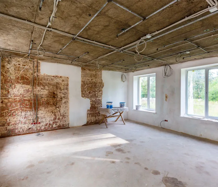 A room under renovation with exposed ceiling wiring, partially stripped walls, two windows, and a small table holding paint buckets.