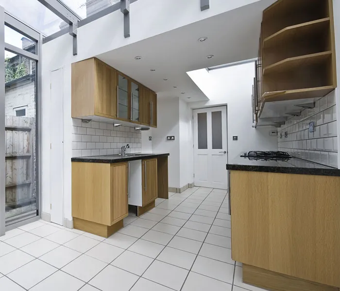 A modern kitchen with wooden cabinets, white tiled walls, black countertops, and a glass door leading outside.