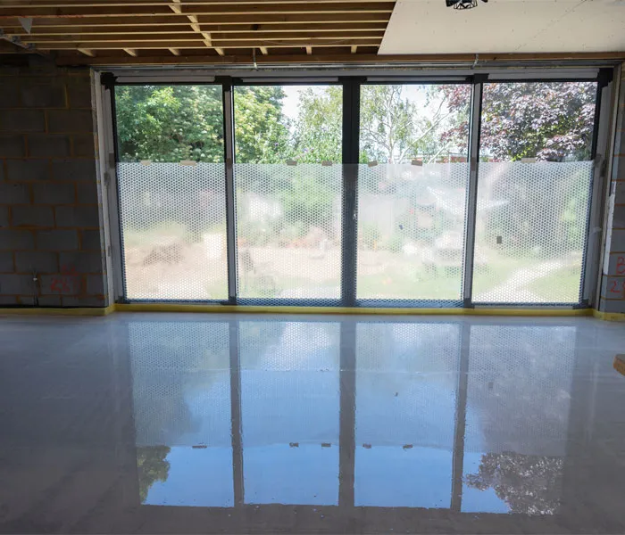 Large floor-to-ceiling windows in an unfinished room overlook a garden. The floor is glossy, and the ceiling framework is exposed.