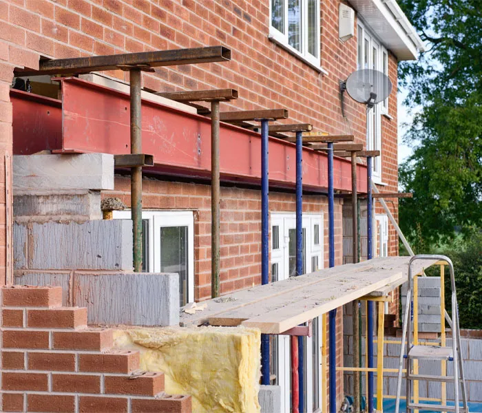 A residential brick building under renovation with scaffolding and steel beams installed above ground-floor windows and doors.