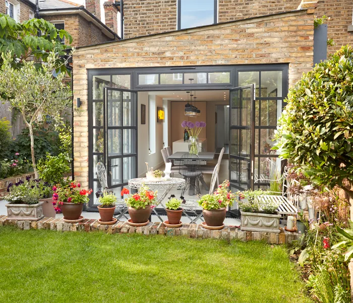 A brick house with open glass double doors leads to a garden featuring a patio with potted plants and outdoor furniture.