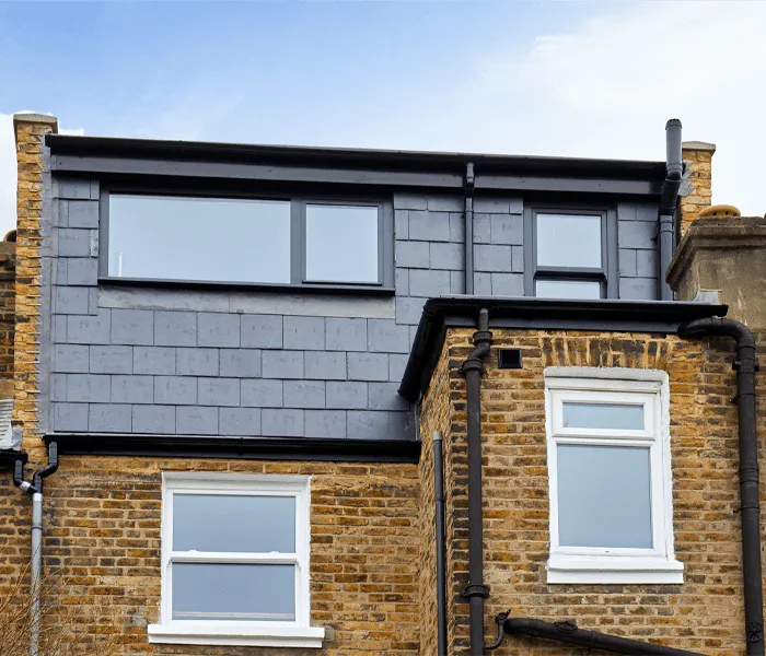 Three-story brick house with dark slate cladding on the top floor and large windows under a blue sky.