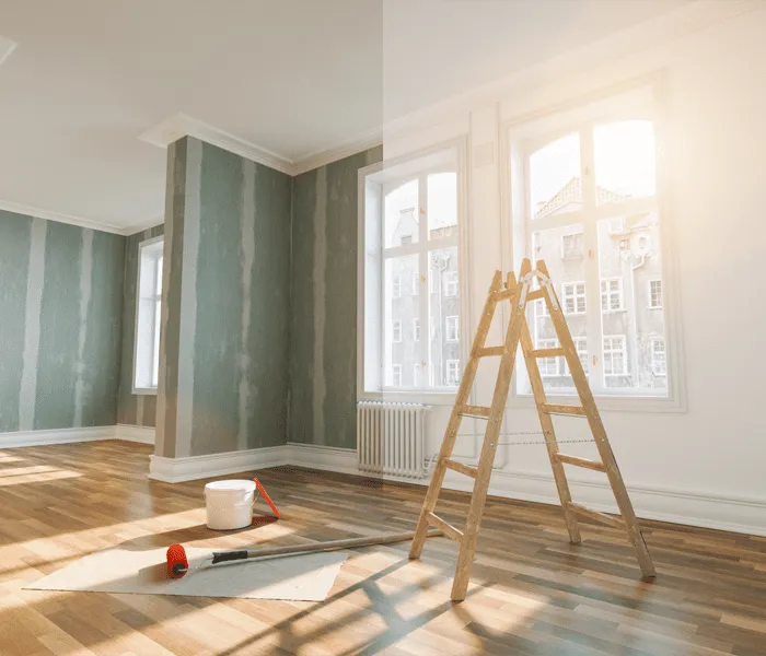 A sunlit room with wooden floors, unpainted walls, a ladder, paint roller, red pan, and white paint bucket in the center.