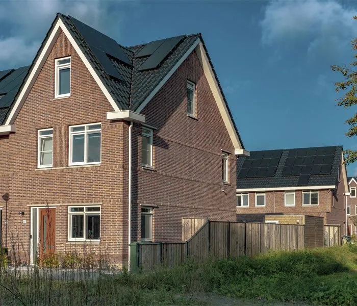 Two modern brick houses with steep roofs and solar panels, surrounded by a wooden fence and greenery under a blue sky.