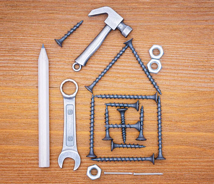 A hammer, pencil, wrench, nuts, nails, and screws arranged on a wooden surface to form a house shape.