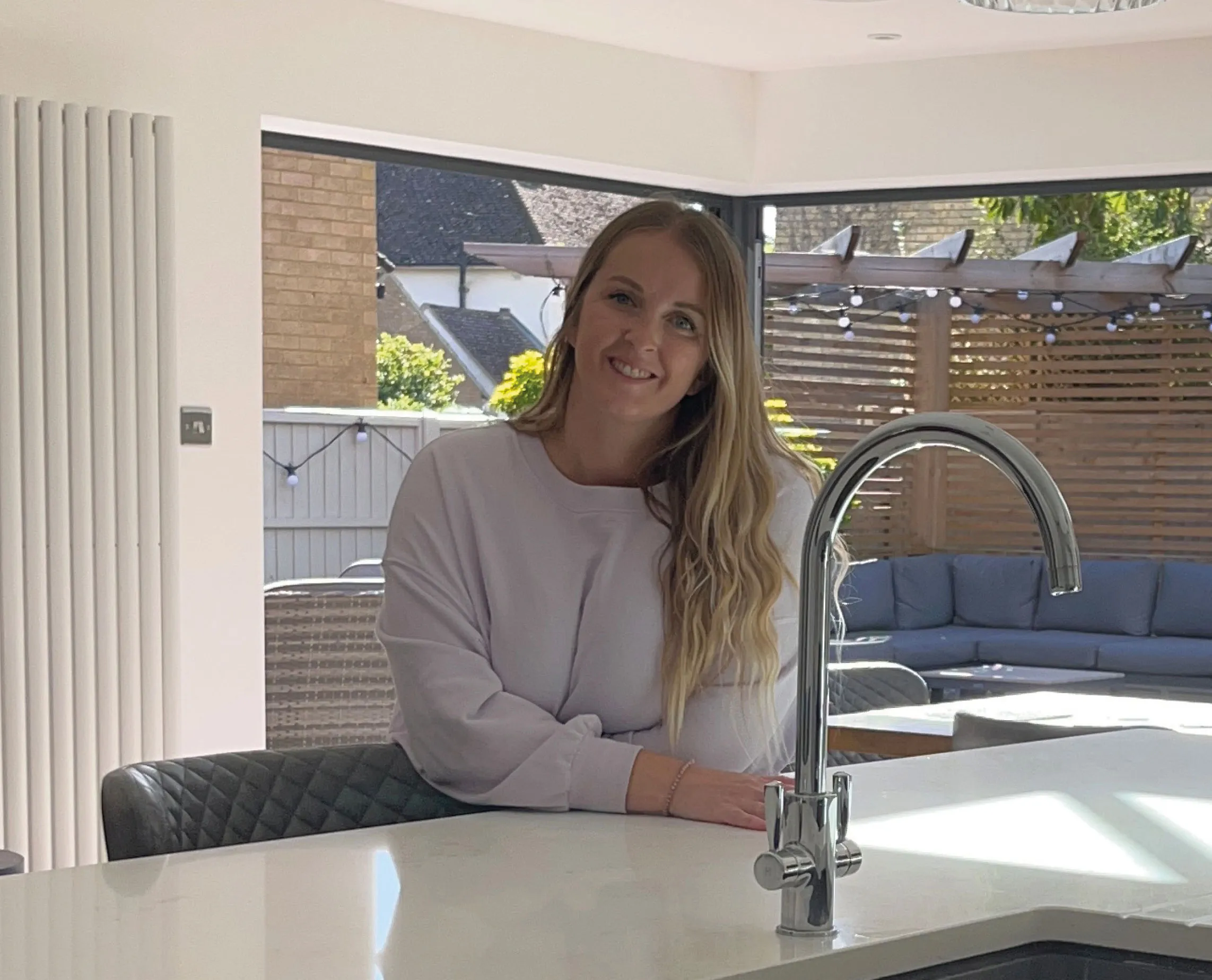 A woman with long blonde hair stands in a modern kitchen by a sink, smiling at the camera, with an outdoor seating area visible in the background.