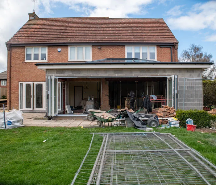 A brick house with a construction site in the backyard, featuring an unfinished extension with debris and building materials scattered around.