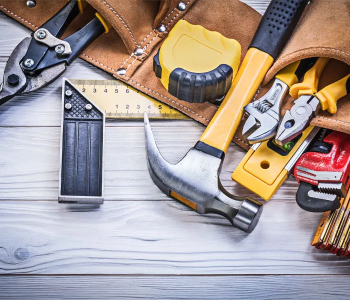 Assorted hand tools, including a hammer, pliers, and measuring tape, arranged on a wooden surface next to a leather tool belt.