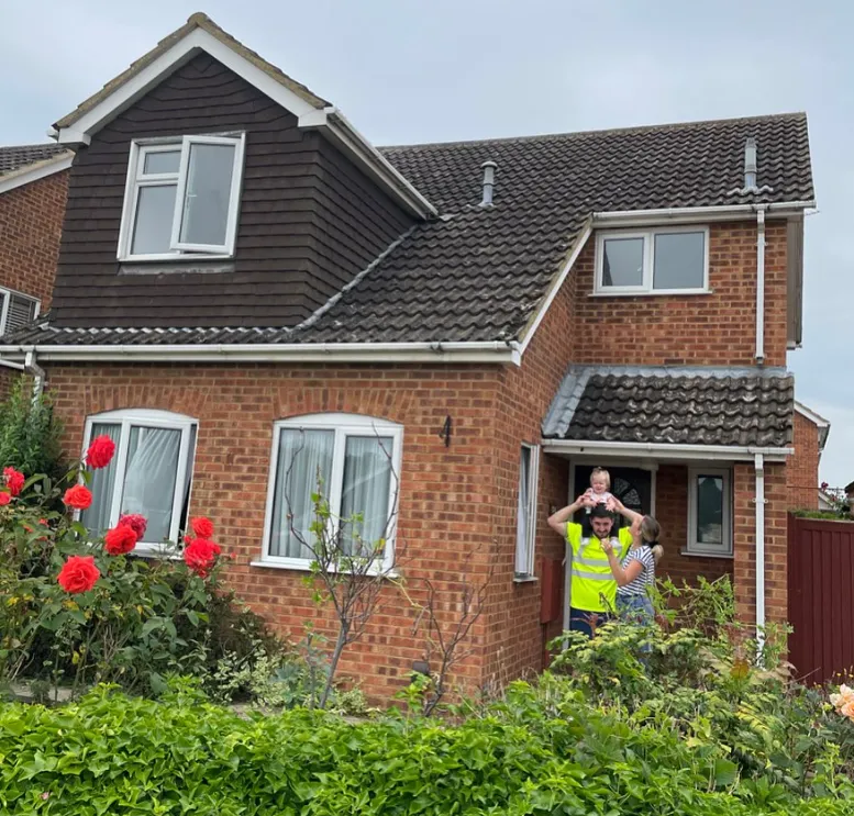 Two people and a child standing in front of a brick house with a garden of red flowers and shrubs.