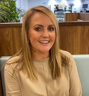 A woman with long blonde hair, wearing a beige sweater, sits in a booth at a cafe with plants and floral decorations in the background.